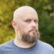 Portrait of a bald, bearded man with blue eyes and nose piercing in a natural outdoor setting.