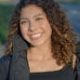Cheerful woman with curly hair and necklace smiling outdoors during the day.